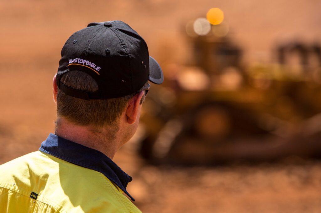 A NPE worker in high-visibility clothing and an “Unstoppable” cap watches heavy machinery operating on a dusty mining site, with the equipment blurred in the background.