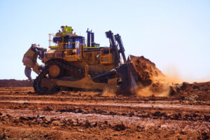 A Caterpillar D11T dozer pushes a heavy load of red earth across a dusty mining worksite under clear blue skies, with dirt and dust flying from the blade as the machine operates.